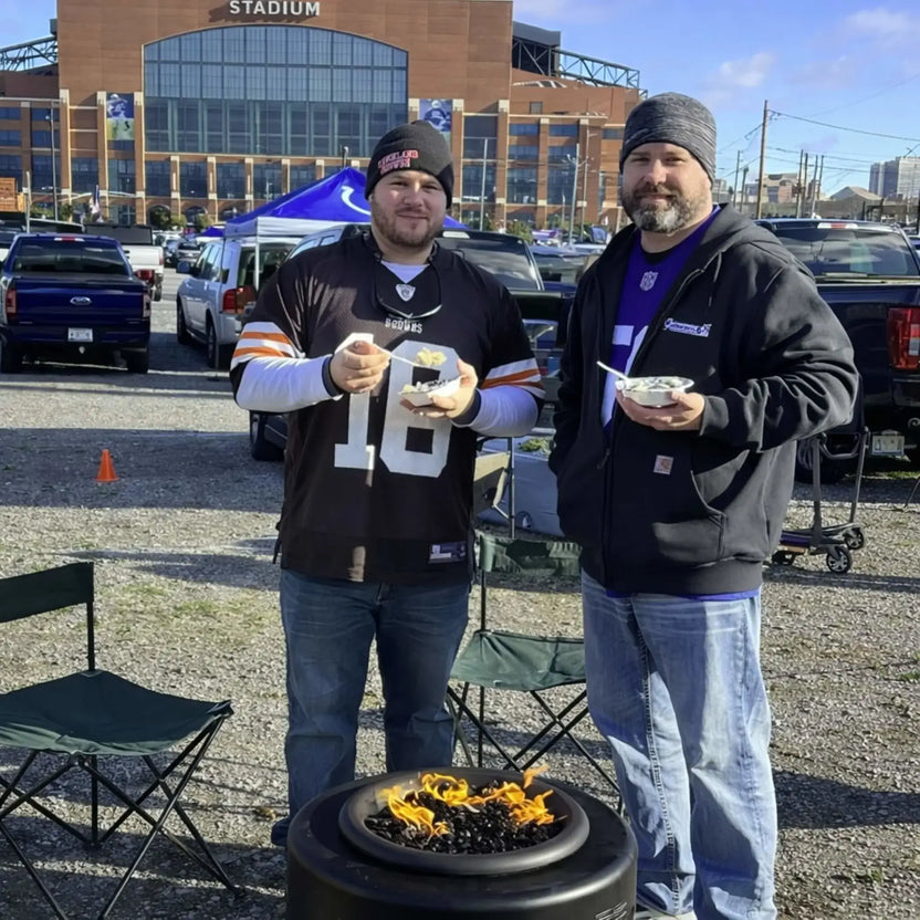 HPC Portable Sport Pit fire pit in a tailgating setup outside a stadium, with two men holding food and standing behind the lit fire pit. The scene highlights portability and social use for sporting events, parking lot gatherings, and outdoor entertaining.