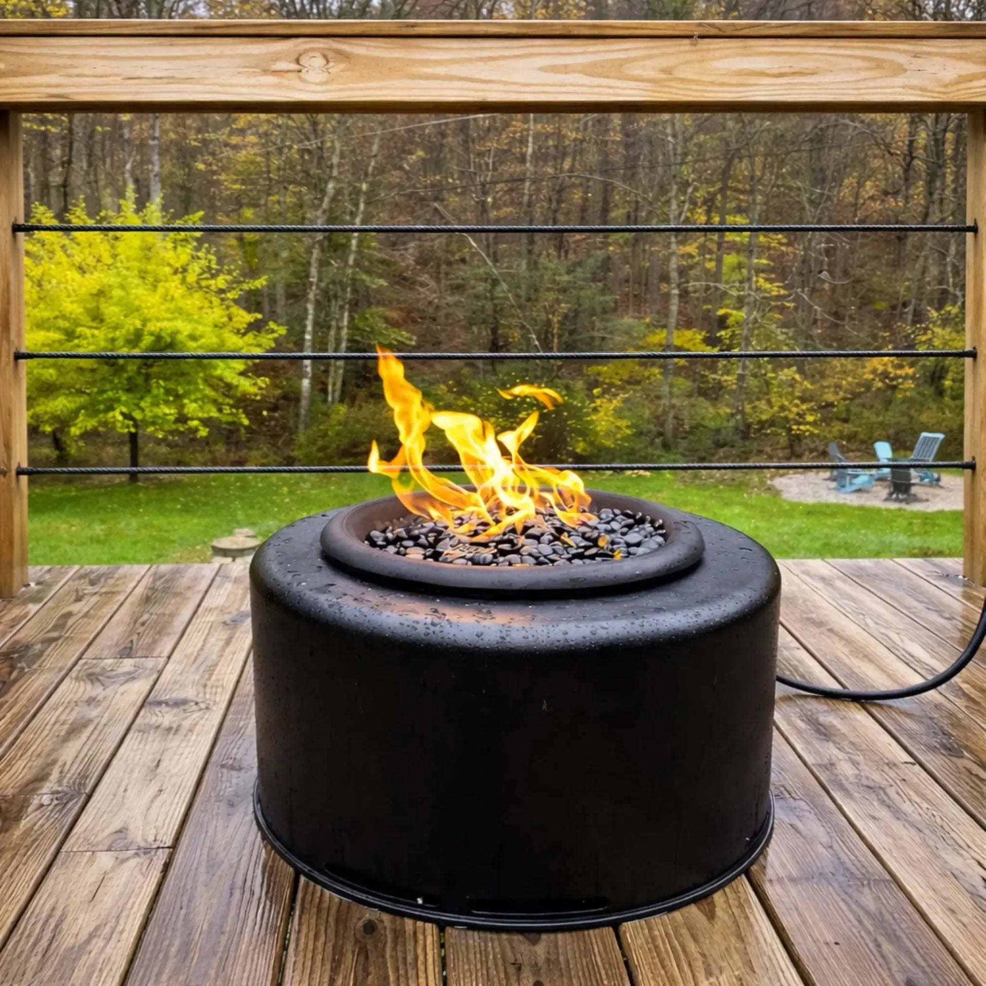 Wide shot of the HPC Portable Sport Pit fire pit centered on a wooden deck overlooking a wooded backyard. Flames rise steadily from black lava rock, with the propane hose visible, emphasizing its portable design and easy outdoor setup.