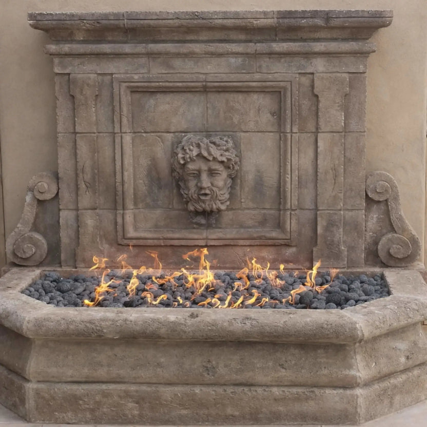 Elegant stone wall fountain with a rectangular fire feature at the base, filled with dark lava rock and bright natural gas flames. The integrated burner creates a clean linear flame pattern, complementing the decorative carved face and rustic masonry finish in an upscale outdoor setting.