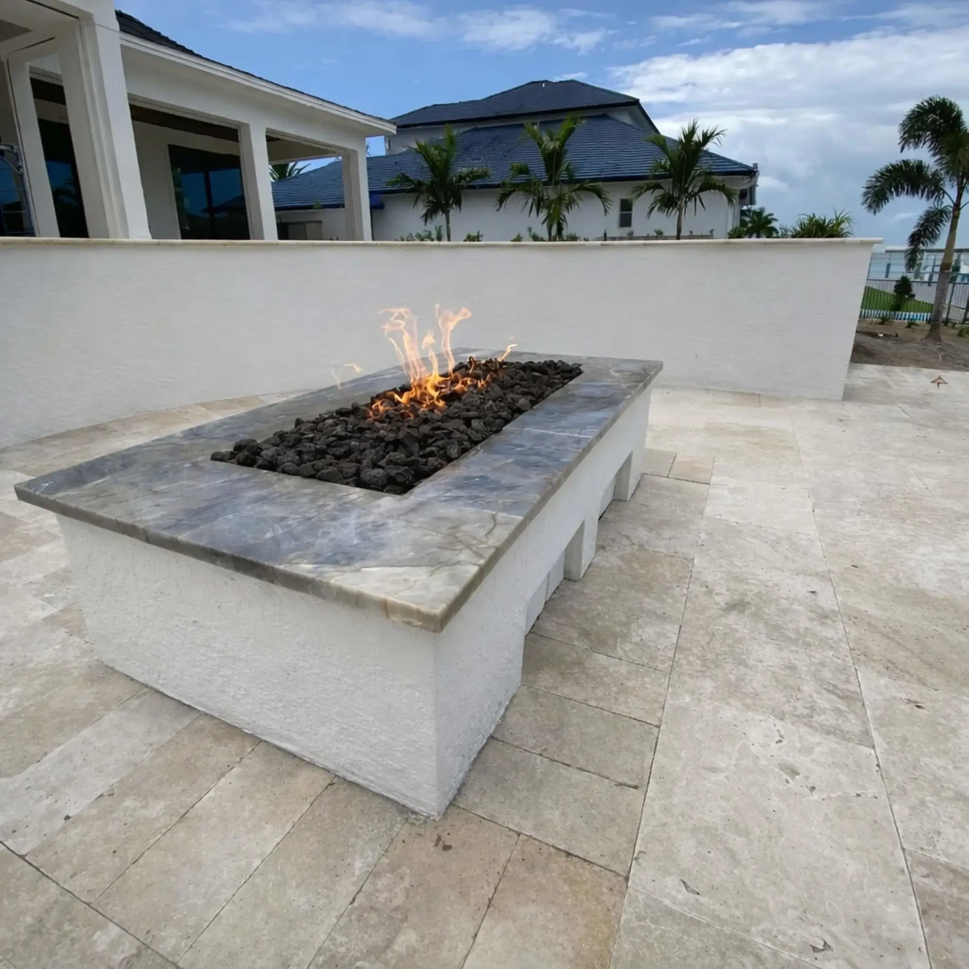 Modern rectangular fire pit with a marble-look top and black lava rock, installed on a spacious tiled patio. The clean flame line contrasts with the bright outdoor setting, framed by a white wall, palm trees, and a contemporary coastal home.
