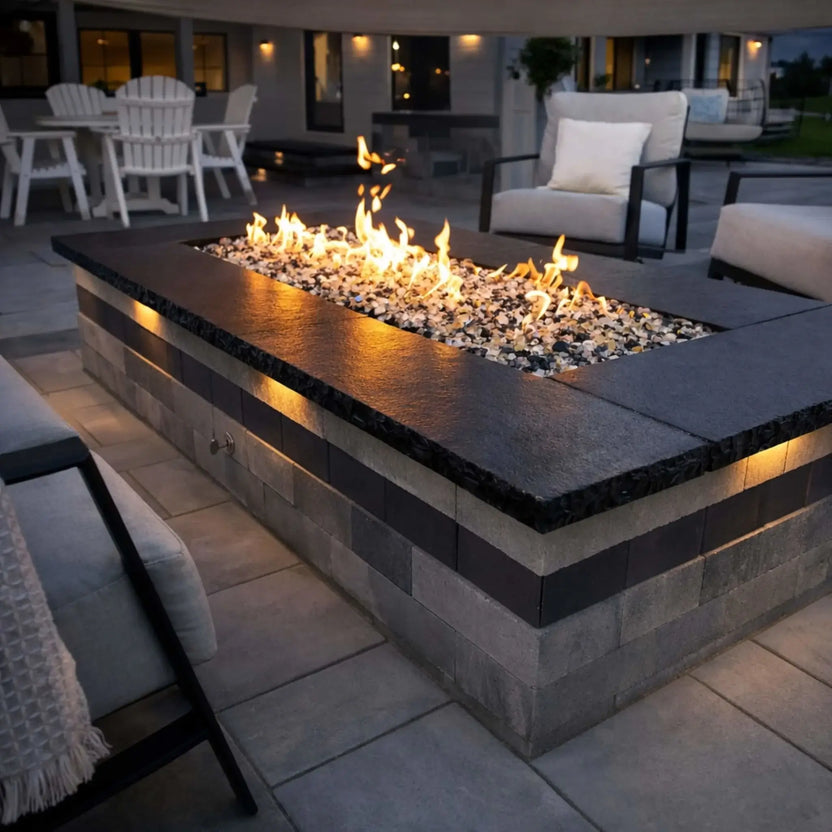 Long rectangular fire feature with a dark stone cap and glass media, producing a continuous linear flame. Set in a covered patio at dusk, the fire pit is surrounded by cushioned seating and ambient lighting, creating a sleek and inviting outdoor lounge.