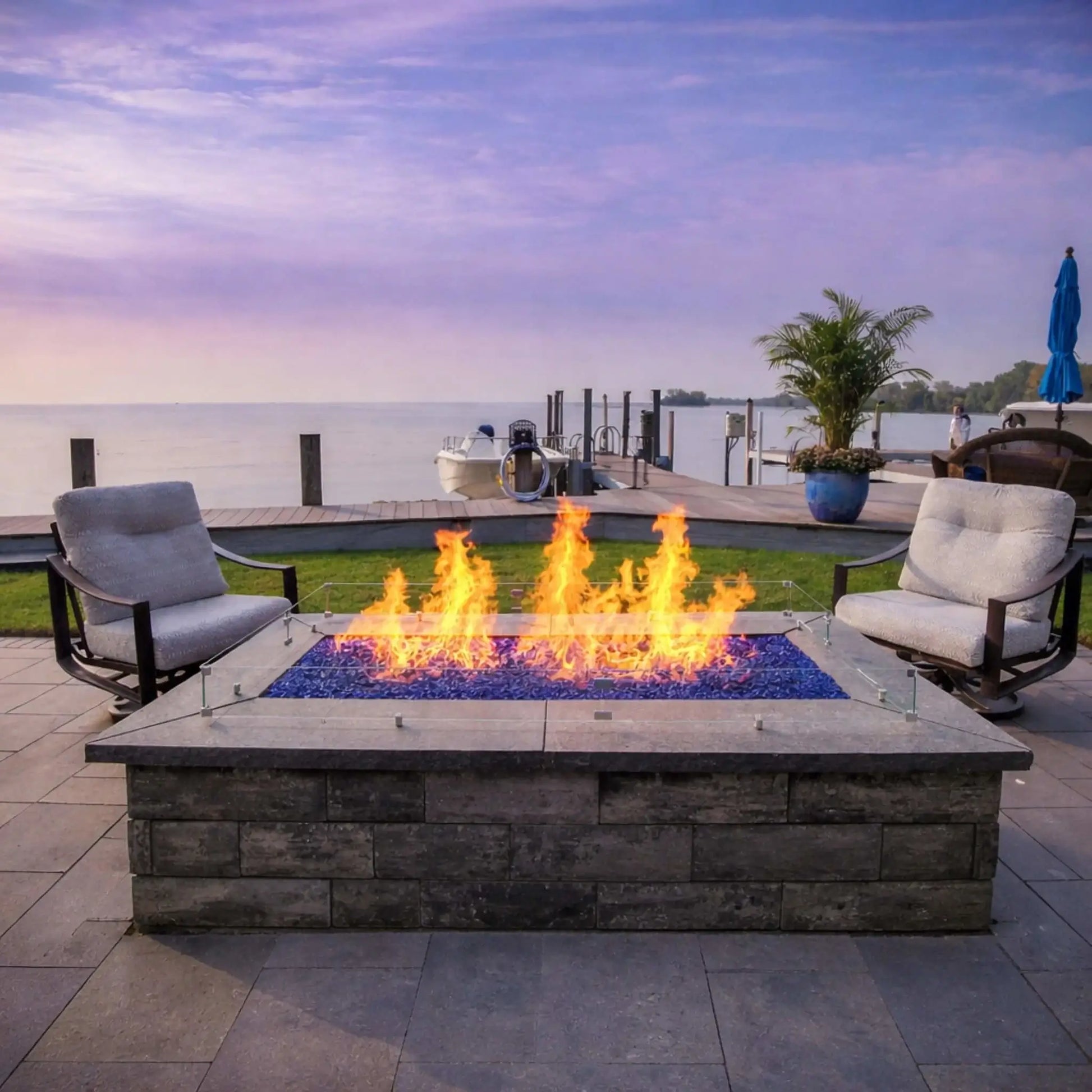 Waterfront patio fire pit with a rectangular burner and blue fire glass, producing tall, vibrant flames. Positioned between two lounge chairs, the fire feature overlooks a calm lake and dock, offering a relaxing, scenic outdoor retreat at sunset.