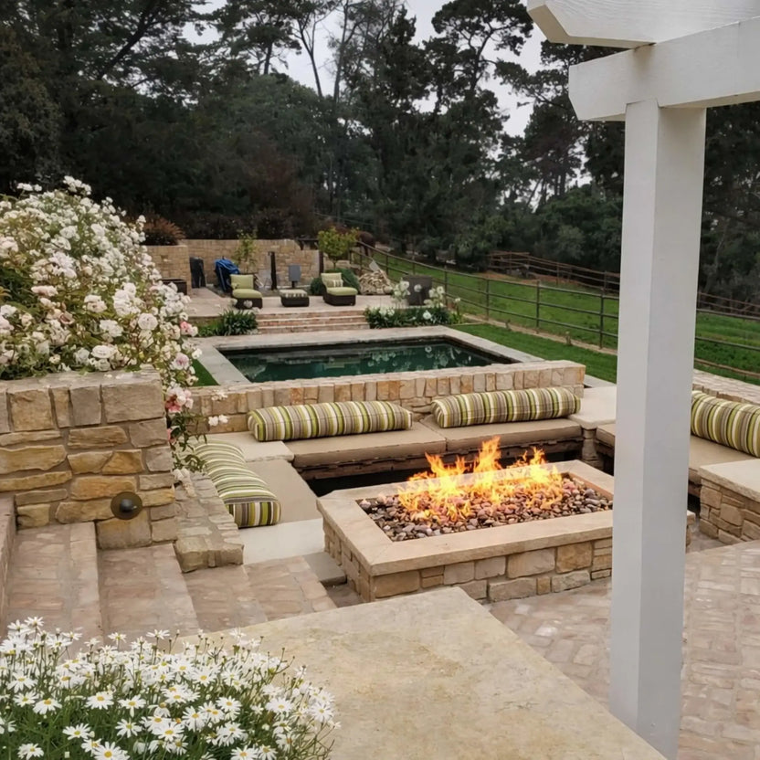 Backyard landscape featuring a rectangular stone fire pit surrounded by built-in seating with striped cushions. Set near a pool and garden area, the fire feature adds warmth and structure to the layered outdoor design with natural stone and greenery.