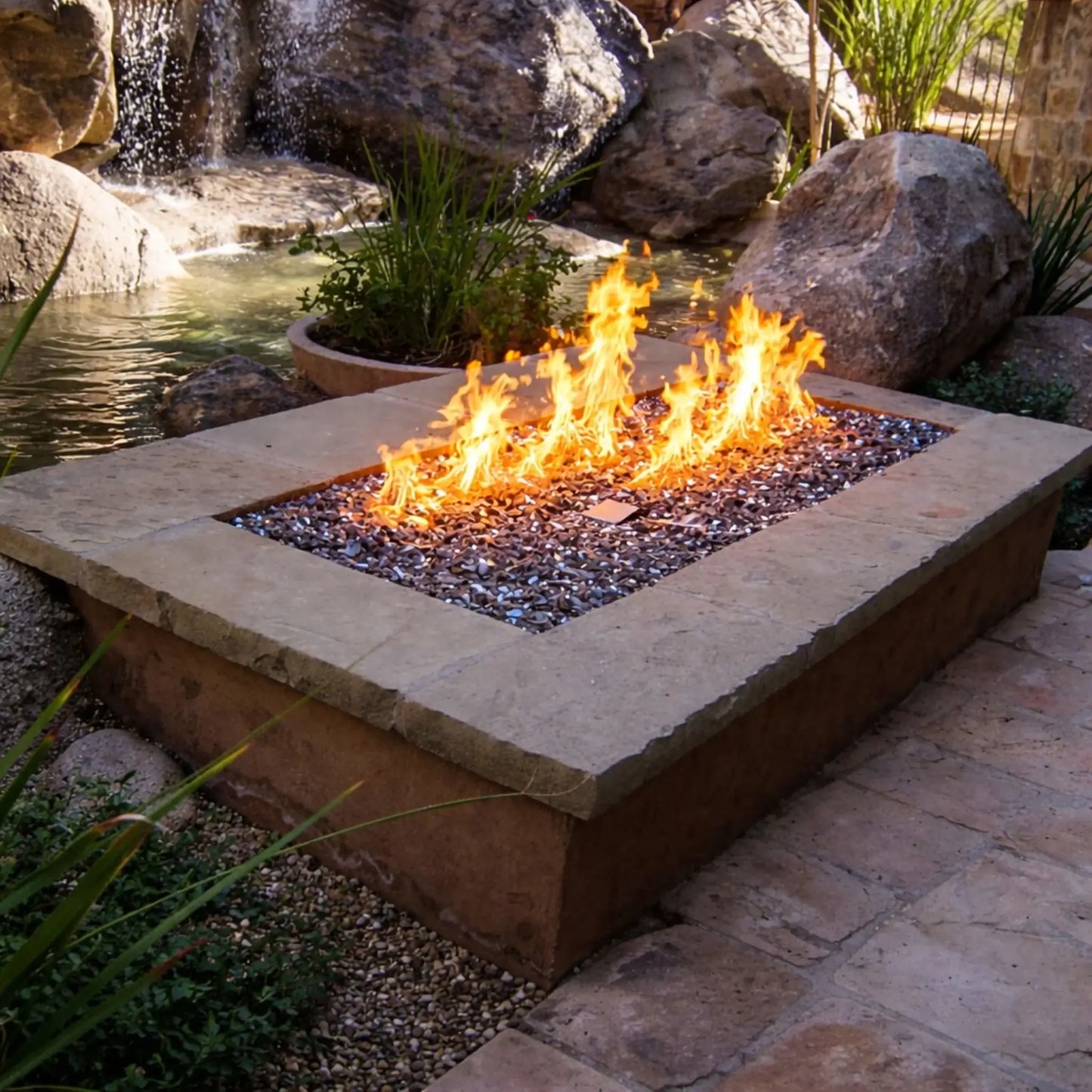 Rustic outdoor fire pit with a rectangular burner and glass media, set beside a natural rock waterfall and pond. Bright flames rise against the backdrop of flowing water, large boulders, and lush plants, creating a tranquil and visually dynamic backyard setting.