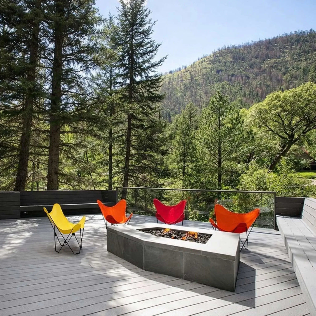 Sleek outdoor deck featuring a built-in rectangular fire pit with a linear burner and glass media, surrounded by colorful lounge chairs. Set against a scenic mountain backdrop, the fire feature provides warmth and visual contrast in a modern elevated patio design.