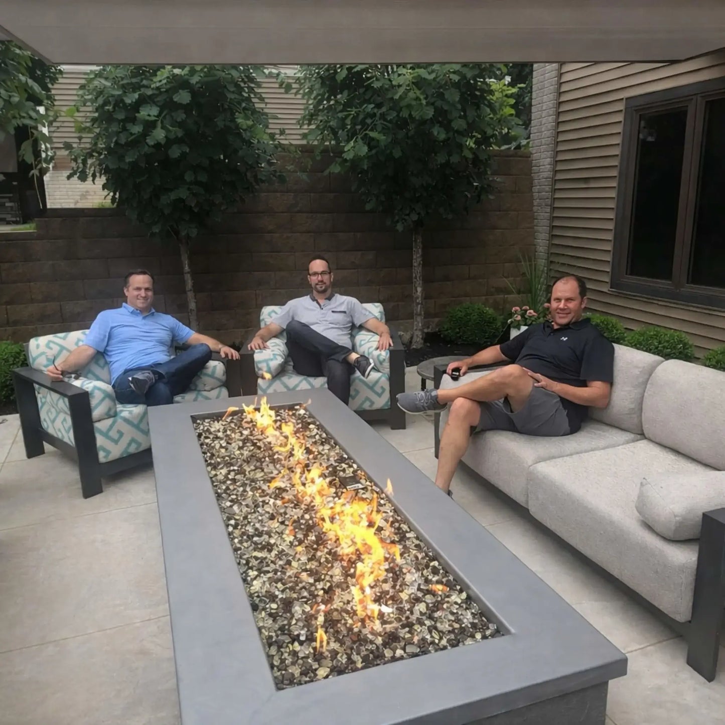 Three men relaxing on cushioned outdoor patio seating gathered around a modern rectangular fire table with a linear gas burner. The fire glass media glows beneath steady flames, creating a warm, inviting setting surrounded by privacy walls and landscaped greenery in a residential backyard.