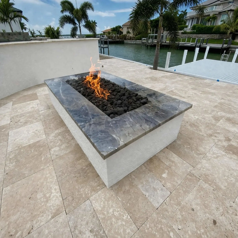 Rectangular fire pit with a polished stone top and black lava rock media, set on a waterfront patio overlooking a canal lined with palm trees and luxury homes. The centered flame adds a subtle focal point to the clean, coastal outdoor living space.