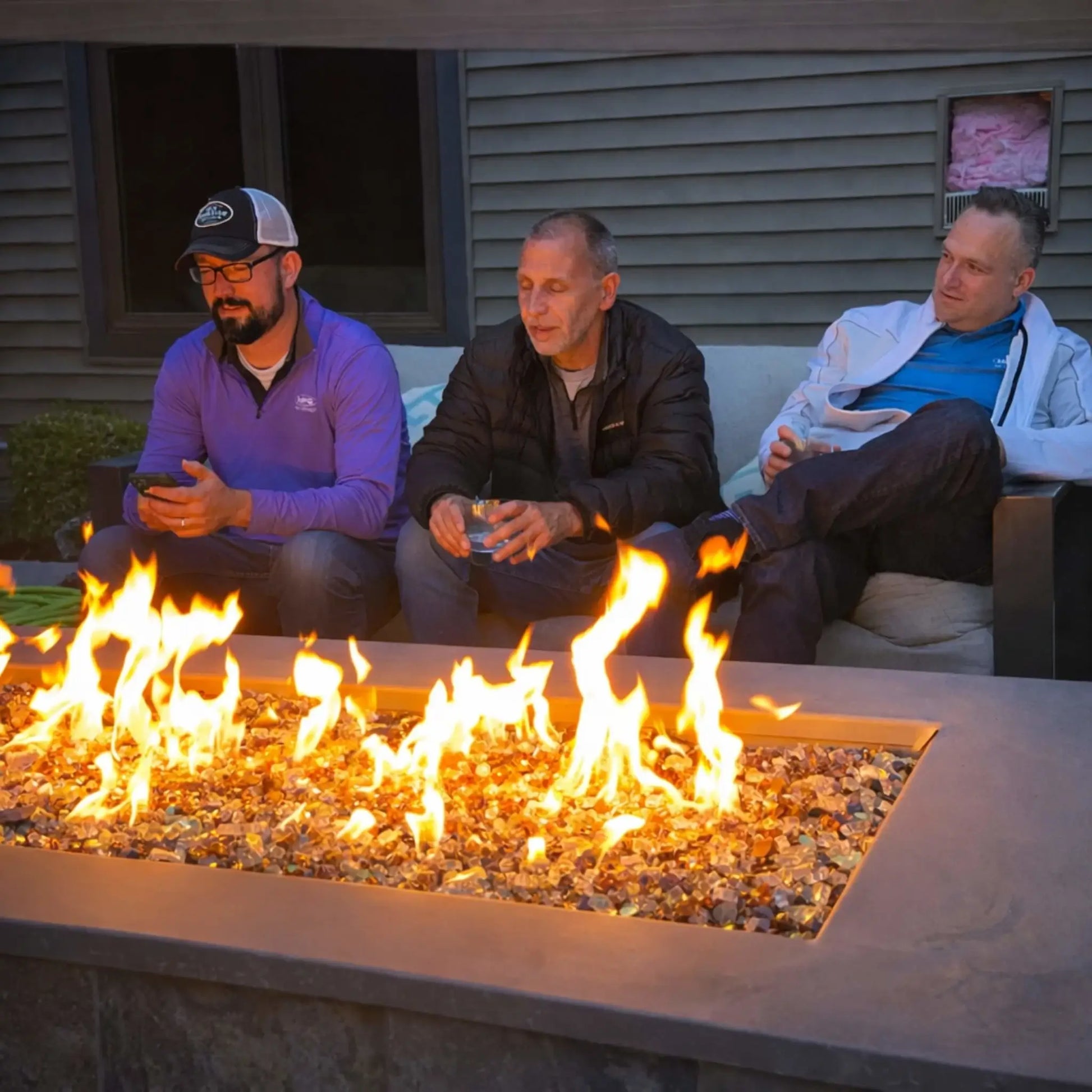 Three men sitting close around a rectangular fire table at dusk, enjoying drinks and conversation. Bright flames rise from reflective fire glass, casting warm light across the seating area and highlighting the cozy, social atmosphere of the outdoor patio.