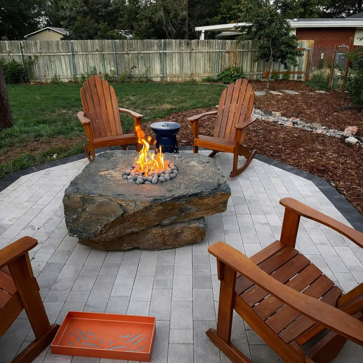 Natural stone fire pit table with rounded lava stones and bright flame centerpiece, surrounded by wooden Adirondack chairs on a circular paver patio. The backyard setting features a wood privacy fence, green lawn, and mulch landscaping, creating a cozy outdoor seating area designed for relaxation and casual gatherings.