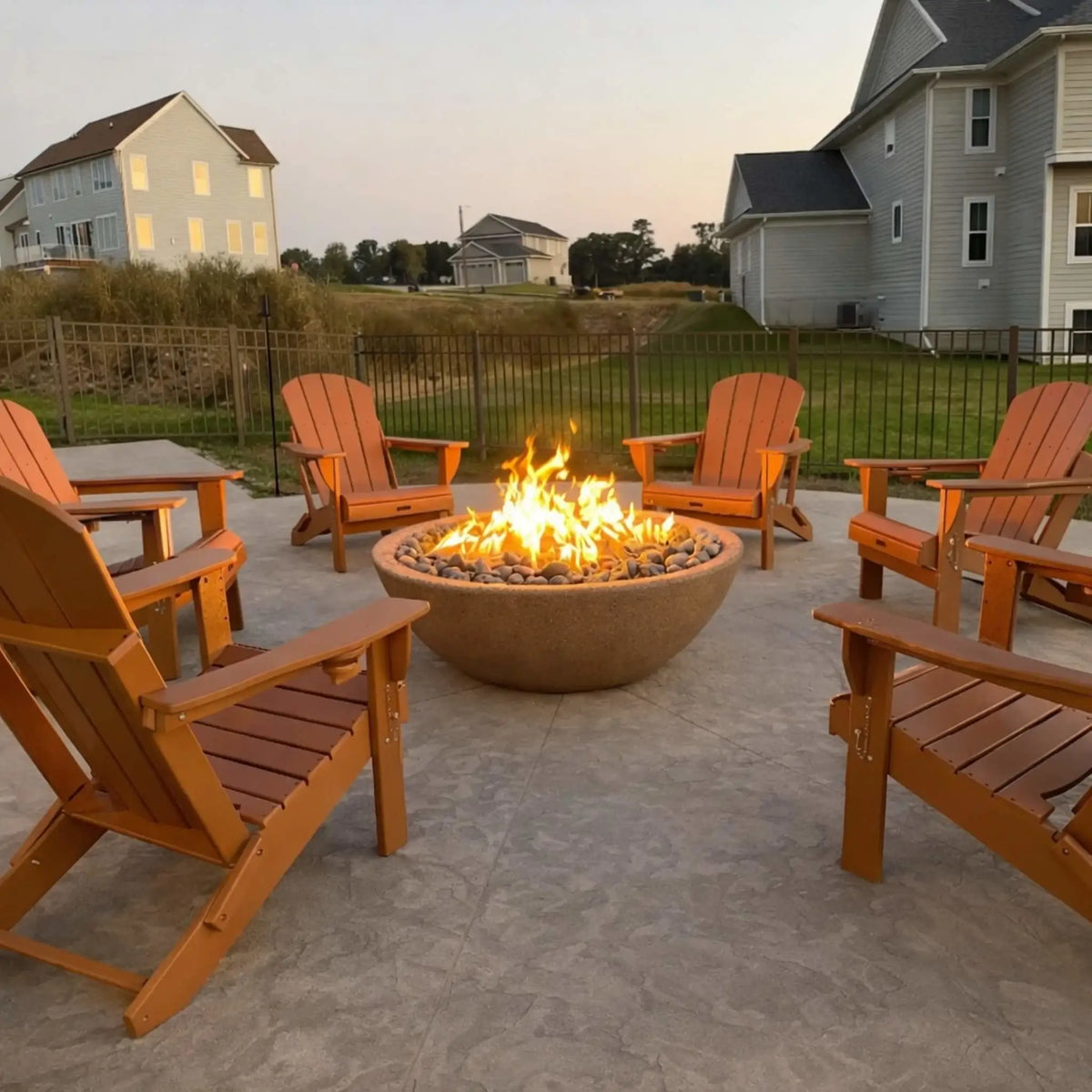 Round gas fire pit filled with smooth lava stones and a bright flame, centered on a stamped concrete patio. Four wooden Adirondack chairs are arranged in a circle, creating a welcoming gathering space in a suburban backyard with neighboring homes, fencing, and open grassy areas at sunset.