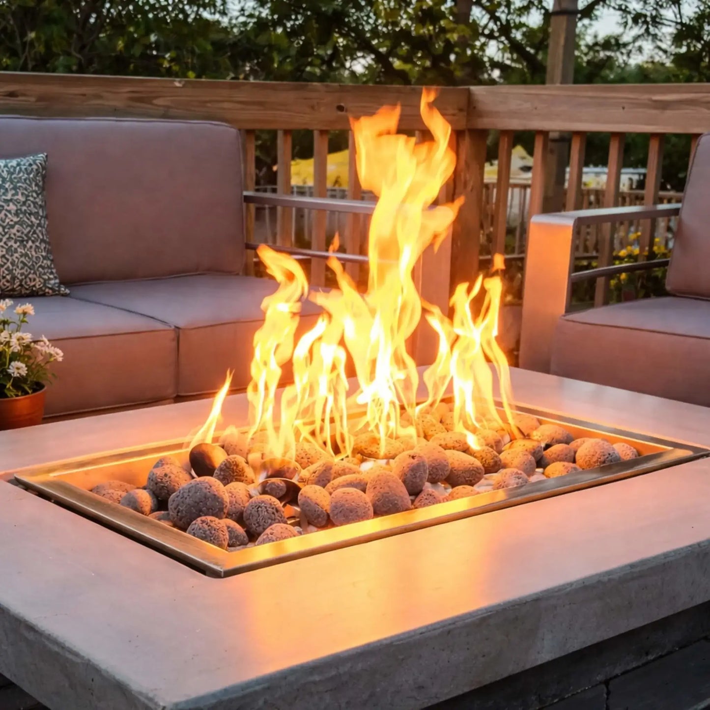 Close-up of a rectangular gas fire table with stainless steel burner tray and rounded lava rocks, producing tall, flickering flames. The setup sits on a wooden deck with cushioned outdoor seating, a small plant, and railing in the background, offering a cozy, modern outdoor lounge feel.