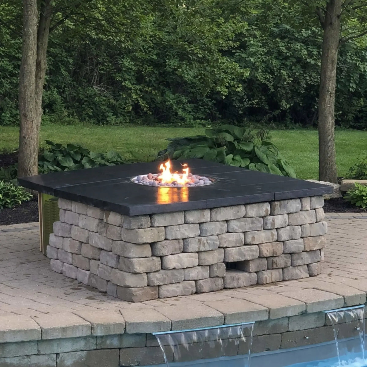 Square stone fire pit with a smooth dark cap and lava rock media, set on a paver patio beside a pool with a small waterfall feature. Surrounded by trees and greenery, the design blends natural landscaping with a functional fire feature for a relaxing backyard retreat.