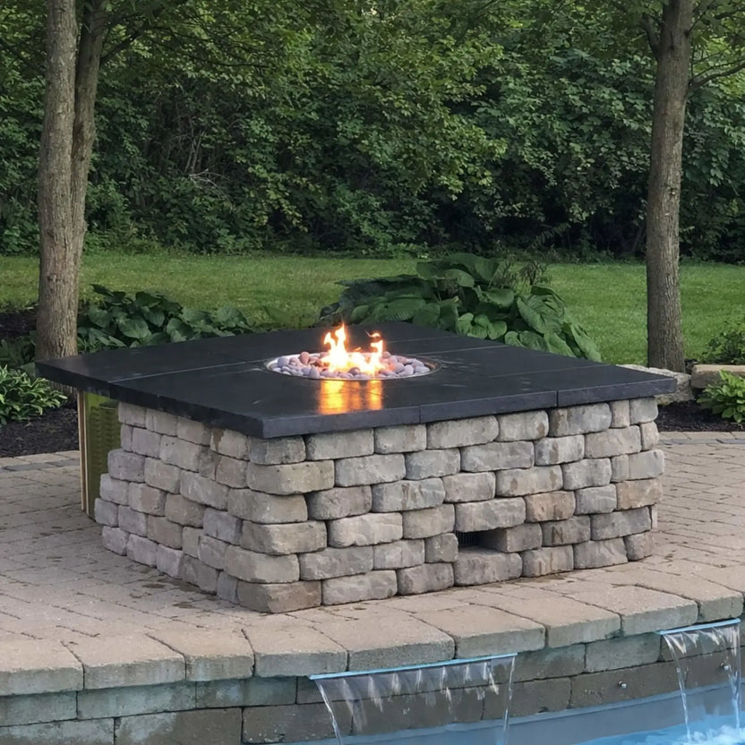 Square stone fire pit with a smooth dark cap and lava rock media, set on a paver patio beside a pool with a small waterfall feature. Surrounded by trees and greenery, the design blends natural landscaping with a functional fire feature for a relaxing backyard retreat.