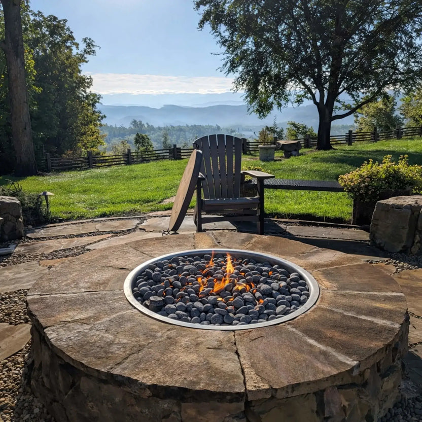 Circular natural stone fire pit filled with dark lava stones and small flames, positioned on a flagstone patio overlooking rolling hills and a scenic mountain view. A single Adirondack chair and rustic fencing enhance the peaceful, countryside setting.