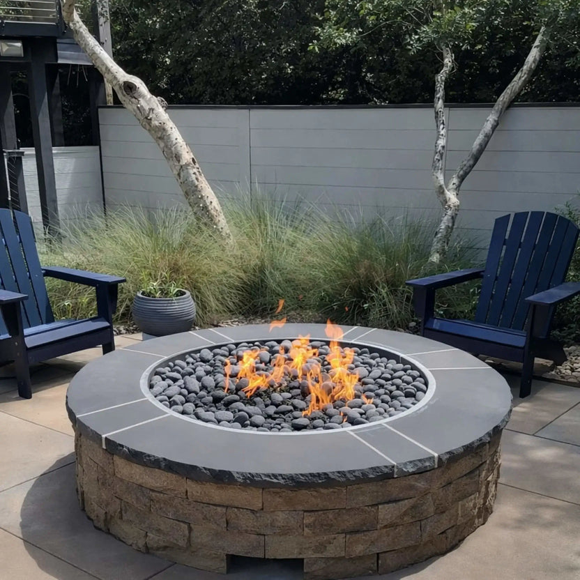 Round gas fire pit with black lava rocks and steady flames, surrounded by blue Adirondack chairs on a clean patio. The landscaped yard includes ornamental grasses, potted plants, and a modern privacy fence, creating a polished and inviting outdoor seating area.