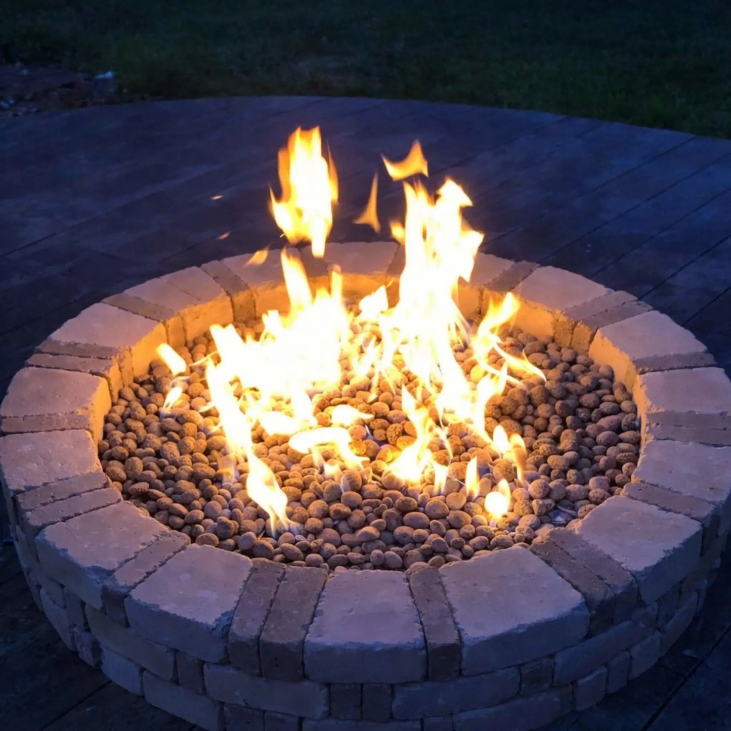 Close-up of a circular stone fire pit filled with lava rock media and bright flames, glowing against a darkened evening backdrop. The stone surround and warm light emphasize the fire’s intensity, making it the focal point of a nighttime outdoor setting.