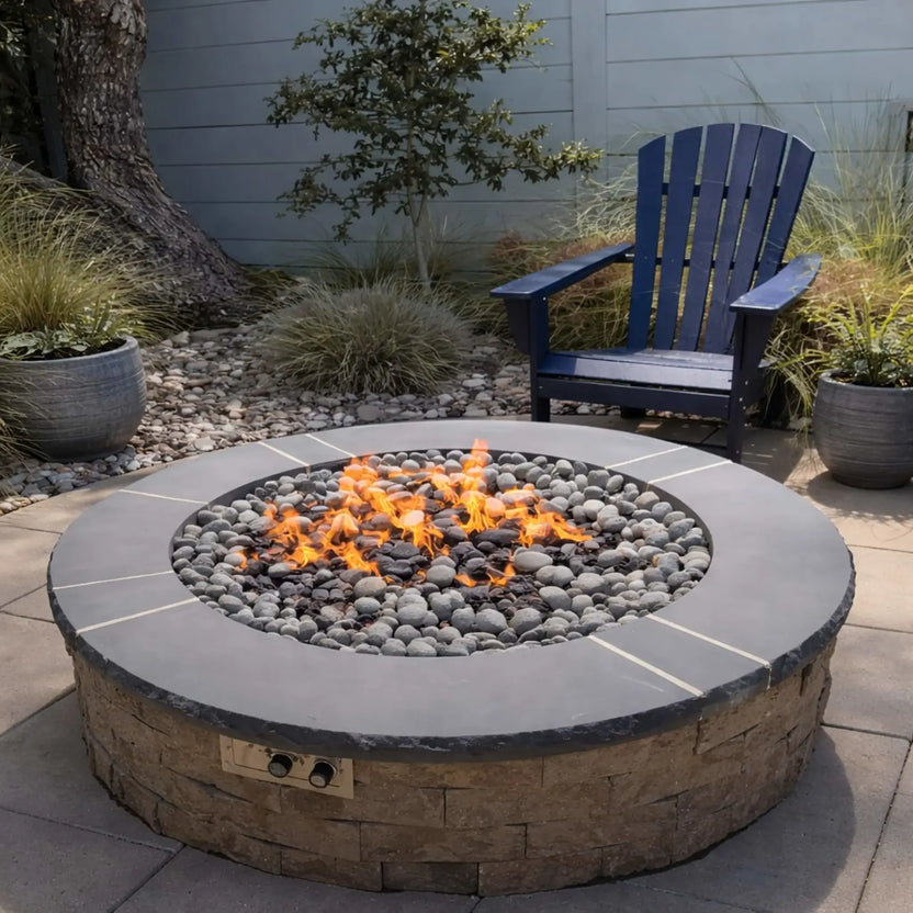 Round gas fire pit with smooth lava stones and active flames, set on a patio near a lakeside yard. Two black outdoor chairs face the fire, while a dock and calm water are visible in the background, creating a relaxing waterfront outdoor living space.