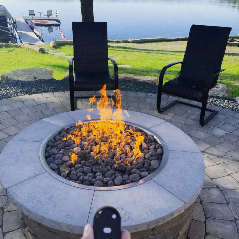 Modern linear gas fire feature with lava rock media set into a sleek black rectangular base, placed on a large tiled patio outside a contemporary home. Expansive windows, clean lines, and built-in seating create a high-end outdoor living environment centered around the fire.