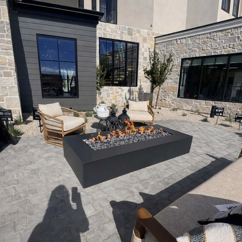 Modern rectangular gas fire pit with black steel finish and lava rock media, producing a clean linear flame on a gray paver patio. The setup sits outside a contemporary home with stone and siding exterior, large black-framed windows, and cushioned outdoor seating, creating a sleek, upscale outdoor living space.