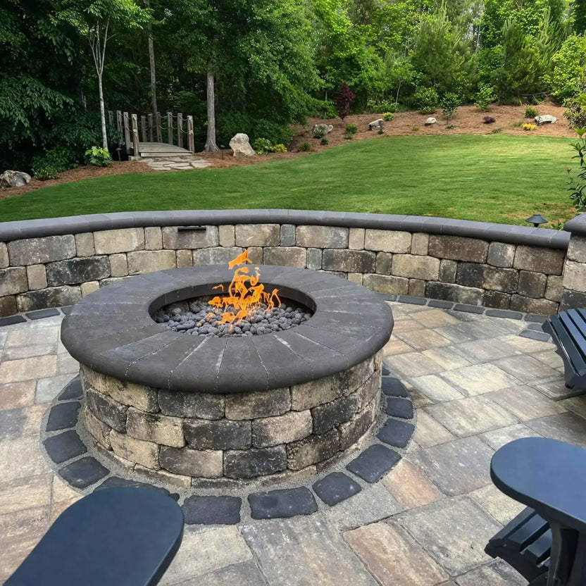 Circular stone fire pit filled with smooth lava rocks and an active flame, built into a paved patio with a matching curved seating wall. The surrounding landscape includes lush green grass, trees, and a small wooden footbridge, giving the space a serene, park-like backyard retreat feel.