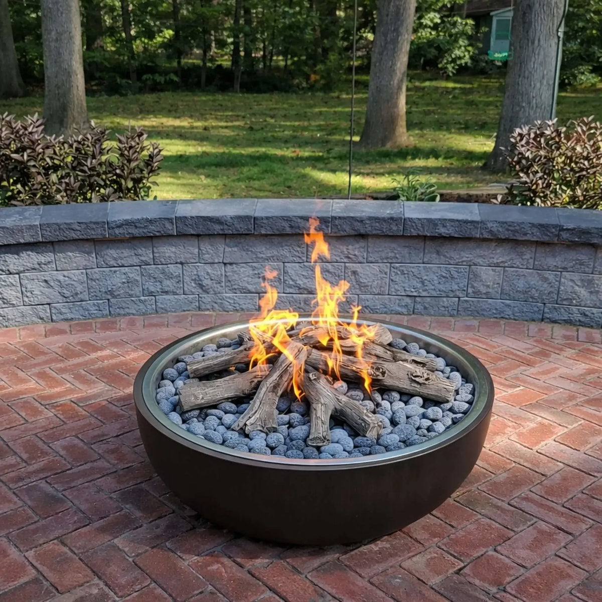Round metal fire pit filled with lava rocks and ceramic log set, burning with natural-looking flames on a brick patio. A curved retaining wall and wooded backyard provide a private, relaxed setting, blending traditional fire pit aesthetics with modern gas functionality.