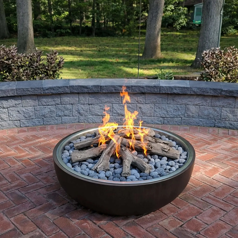 Round metal fire pit filled with lava rocks and ceramic log set, burning with natural-looking flames on a brick patio. A curved retaining wall and wooded backyard provide a private, relaxed setting, blending traditional fire pit aesthetics with modern gas functionality.