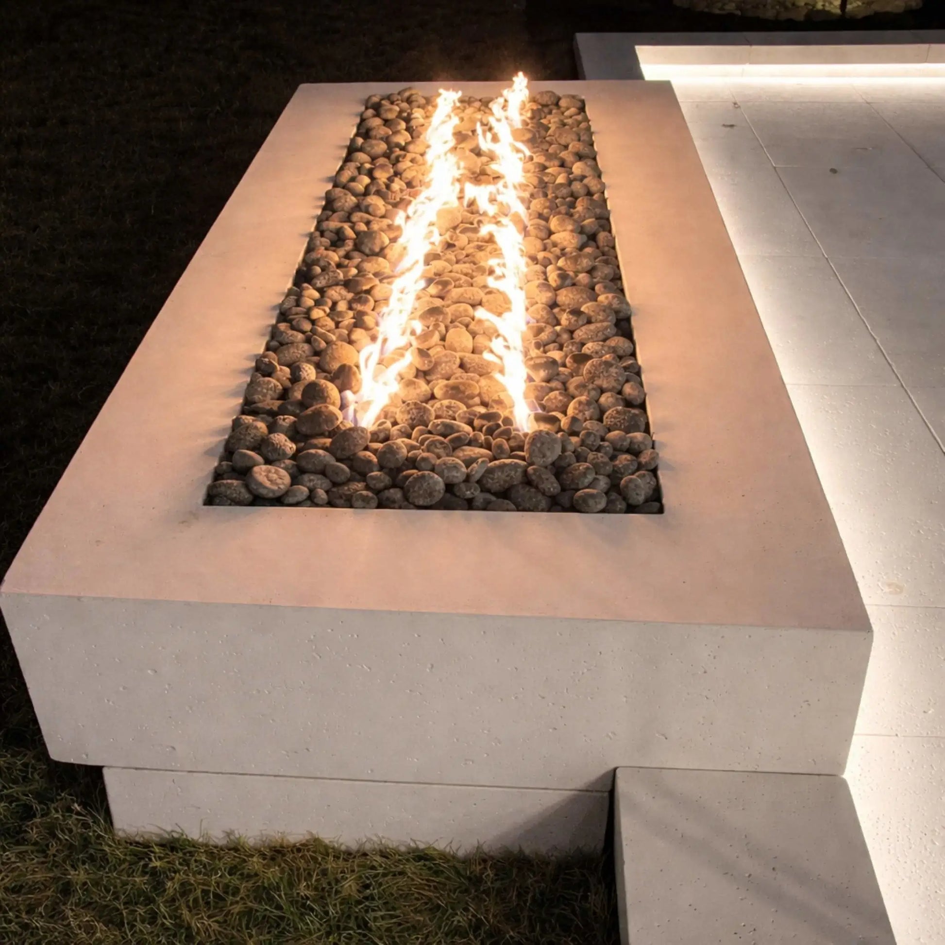 Modern rectangular concrete fire table with a linear burner and polished lava stones, producing tall, even flames. The sleek design sits on a clean patio surface with subtle lighting, emphasizing a contemporary outdoor living space ideal for evening ambiance and entertaining.