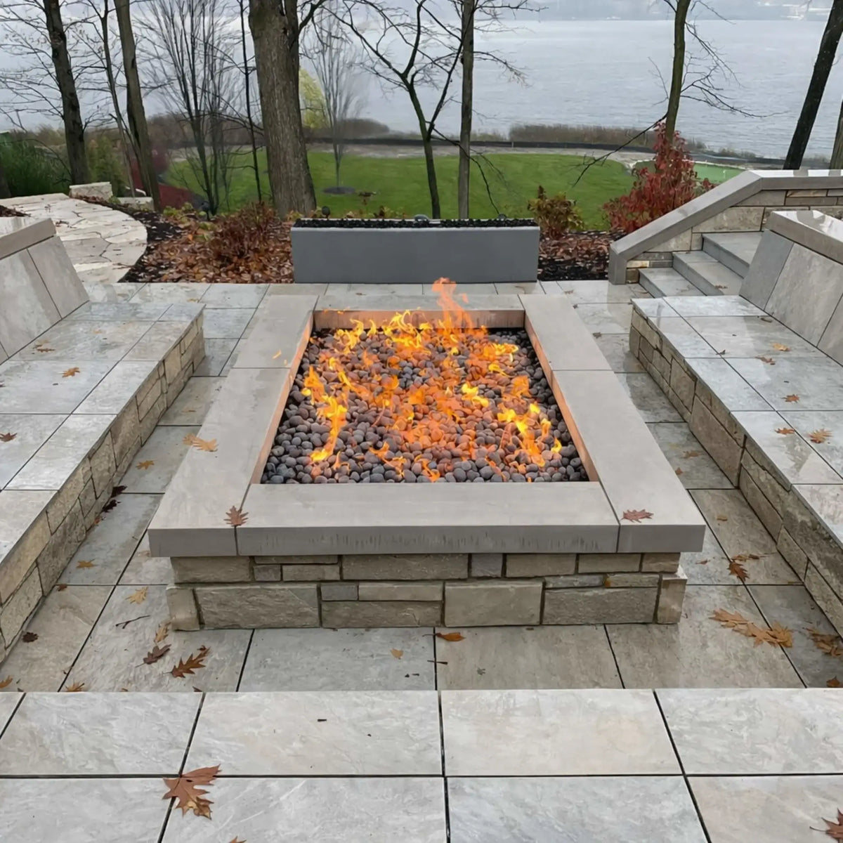 Square gas fire pit filled with dark lava stones and vibrant flames, set within a multi-level stone patio overlooking a lake. Built-in seating walls and surrounding steps frame the fire feature, while fallen leaves and overcast skies create a calm, autumn-inspired outdoor setting.