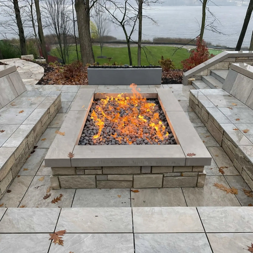 Square gas fire pit filled with dark lava stones and vibrant flames, set within a multi-level stone patio overlooking a lake. Built-in seating walls and surrounding steps frame the fire feature, while fallen leaves and overcast skies create a calm, autumn-inspired outdoor setting.