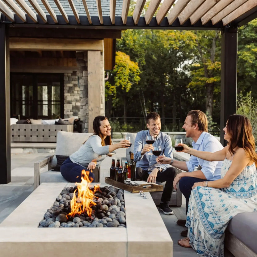Group of friends gathered around a rectangular fire pit table with lava rock media, enjoying drinks under a modern pergola. The outdoor living area features cushioned seating, wood beams overhead, and a warm flame centerpiece that enhances the inviting, social atmosphere.