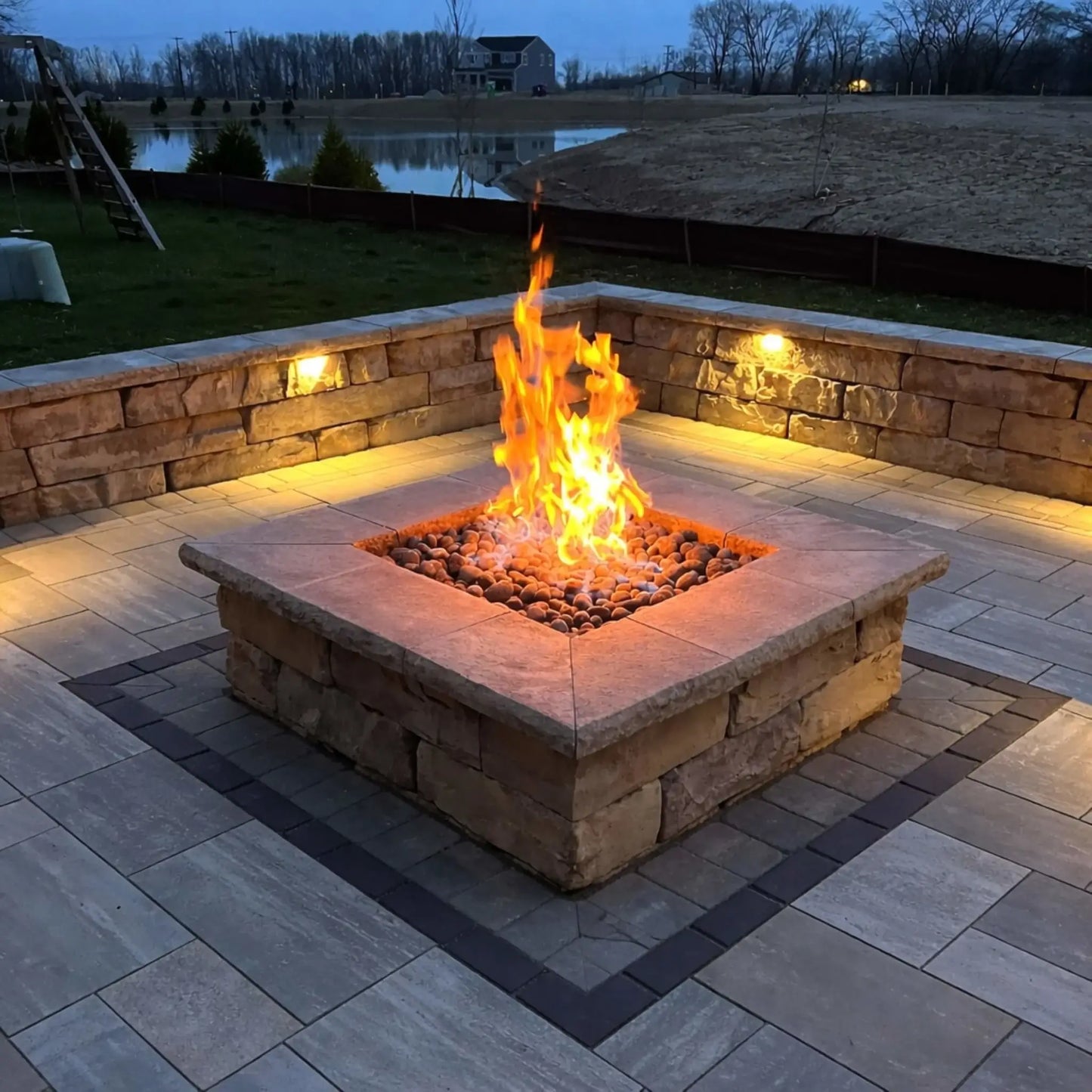 Square stone fire pit with glowing lava rocks and a strong flame, surrounded by a paved patio and illuminated retaining wall. The evening setting overlooks a pond and open landscape, with built-in lighting adding warmth and highlighting the fire feature as a focal point.