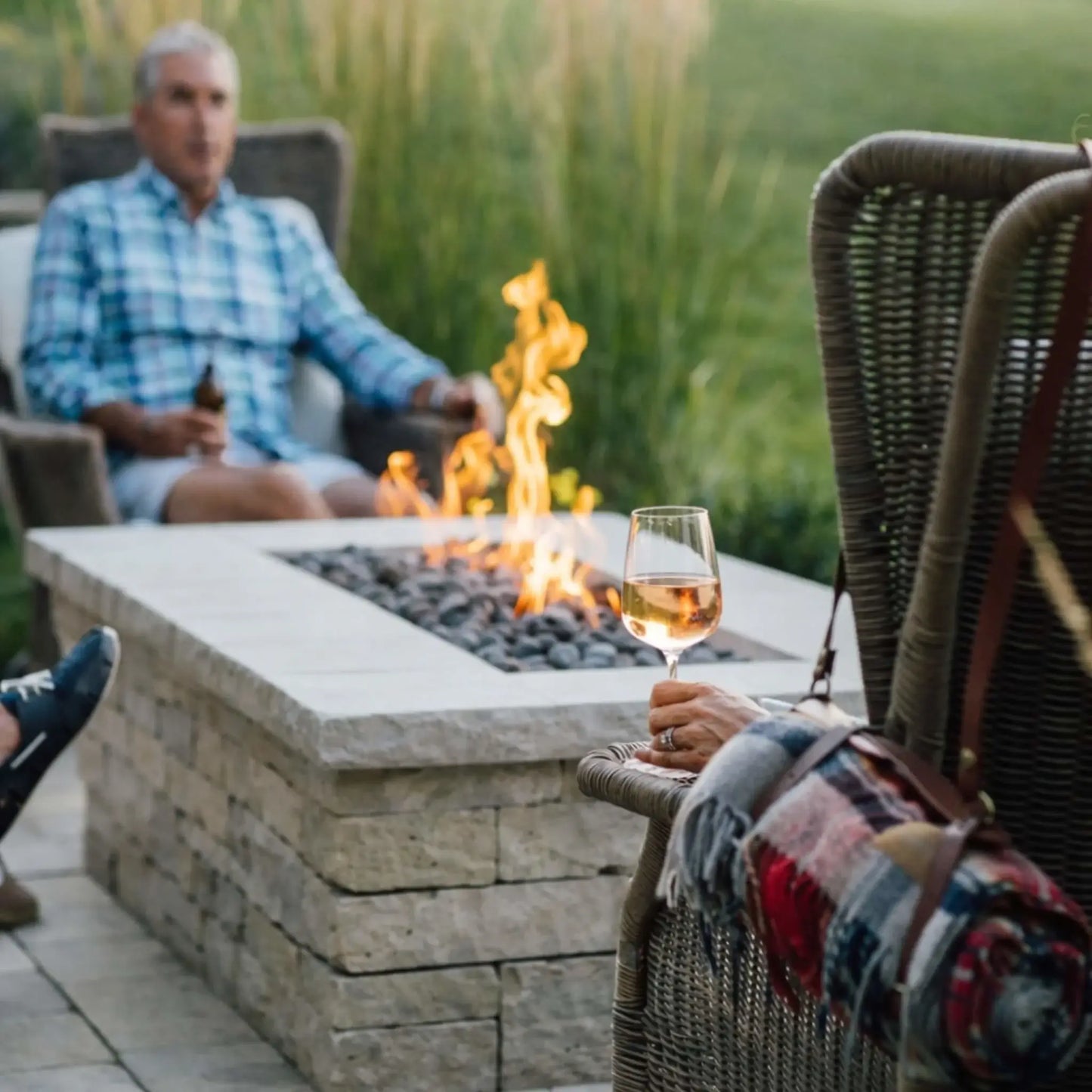 Close-up of a rectangular gas fire table filled with smooth lava stones and steady flames, surrounded by outdoor seating. A person in the foreground holds a glass of wine, while another relaxes in the background, capturing a quiet, intimate backyard moment centered around the fire.