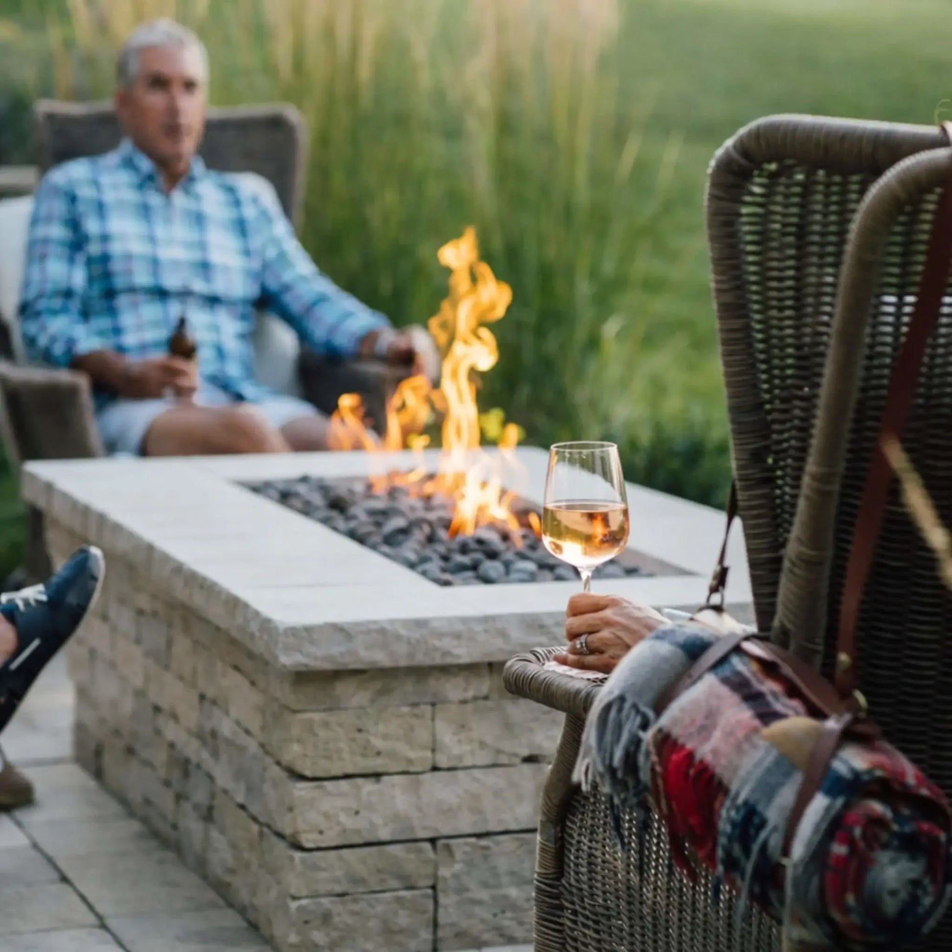 Close-up of a rectangular gas fire table filled with smooth lava stones and steady flames, surrounded by outdoor seating. A person in the foreground holds a glass of wine, while another relaxes in the background, capturing a quiet, intimate backyard moment centered around the fire.