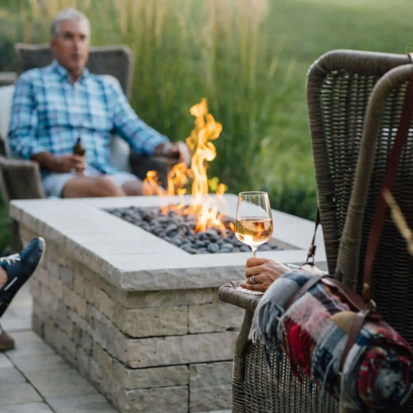 Close-up of a rectangular gas fire table filled with smooth lava stones and steady flames, surrounded by outdoor seating. A person in the foreground holds a glass of wine, while another relaxes in the background, capturing a quiet, intimate backyard moment centered around the fire.