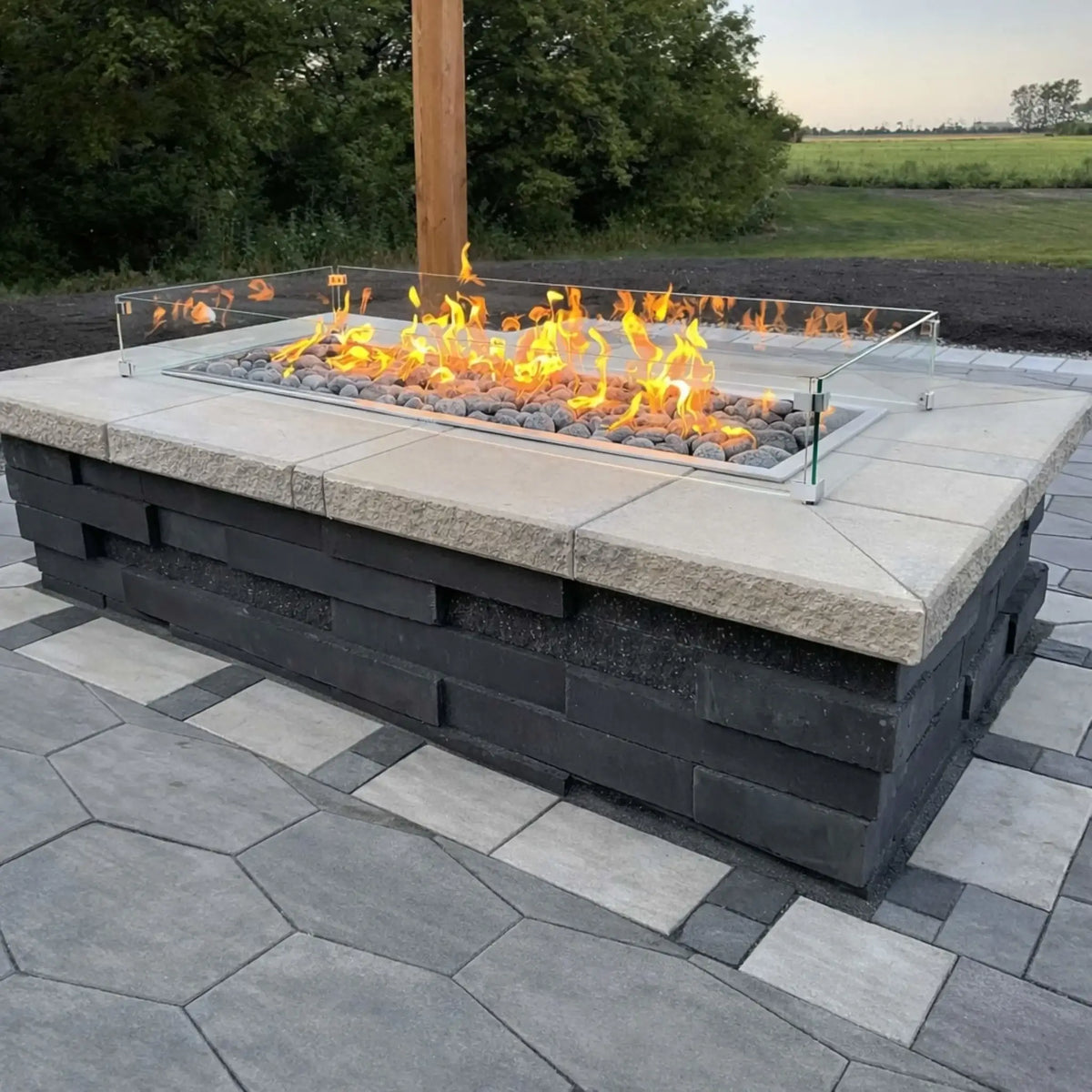 Linear gas fire pit with lava rock media and a glass wind guard, built into a large rectangular patio feature. The flames run evenly across the burner, while the surrounding paver patio and open landscape create a modern, spacious outdoor entertaining area.