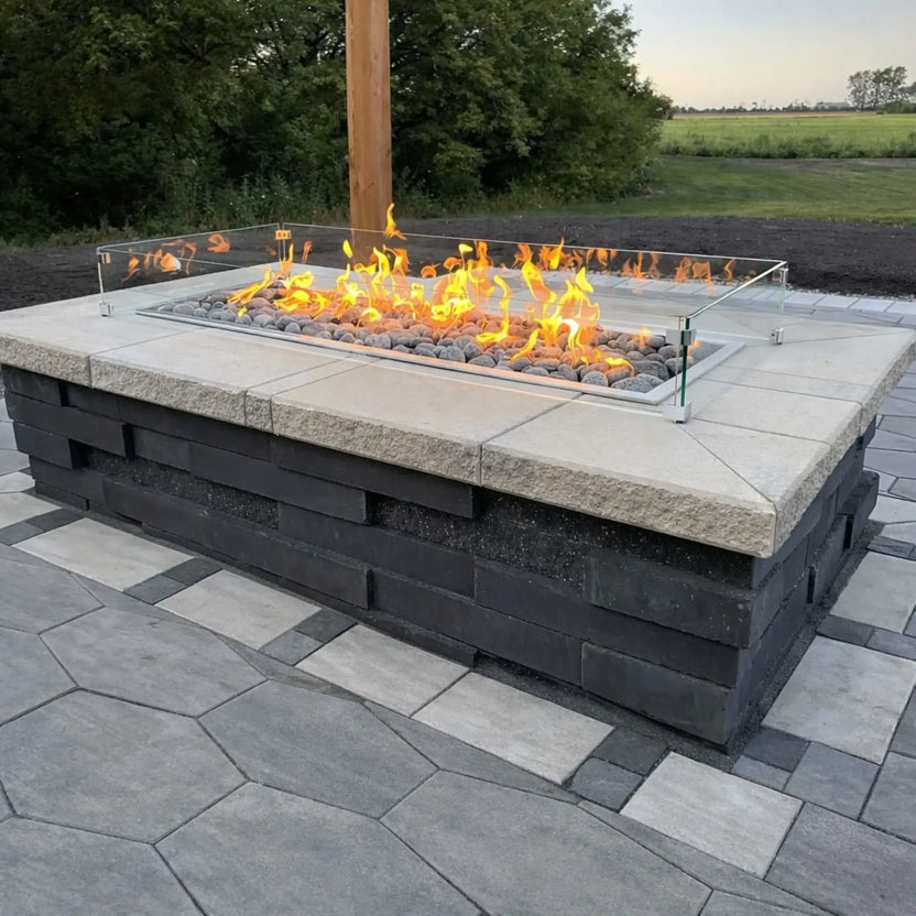 Linear gas fire pit with lava rock media and a glass wind guard, built into a large rectangular patio feature. The flames run evenly across the burner, while the surrounding paver patio and open landscape create a modern, spacious outdoor entertaining area.