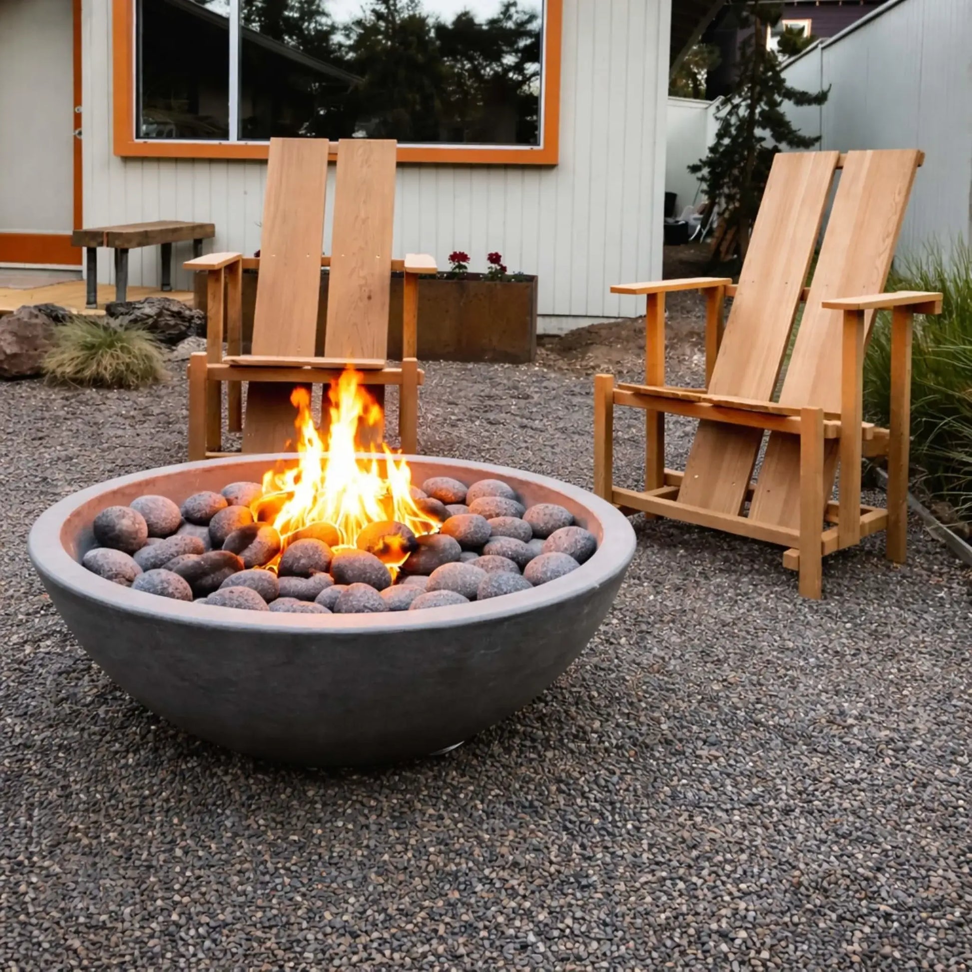 Round bowl-style fire pit filled with smooth lava stones and a lively flame, placed on a gravel patio. Two wooden Adirondack chairs sit nearby, with a modern home exterior in the background, creating a simple yet inviting backyard fire feature setup.