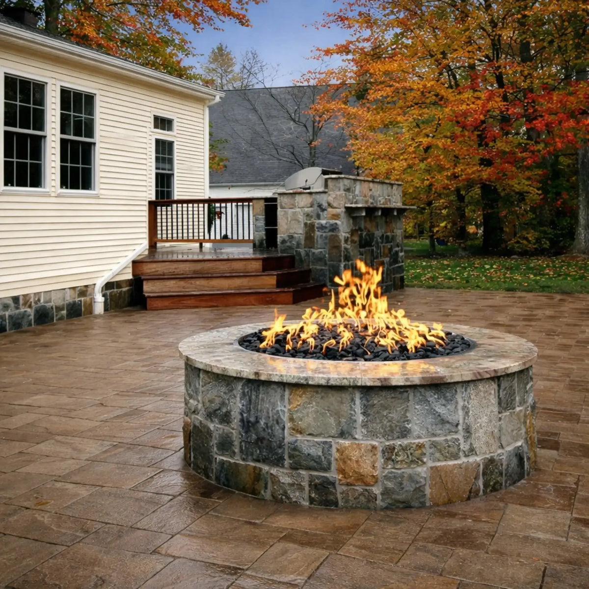 Outdoor backyard patio featuring a circular stone gas fire pit filled with black fire glass, producing a warm flame display. The fire pit sits on a paved surface near a raised deck and outdoor kitchen area, surrounded by autumn trees and a cozy residential setting.