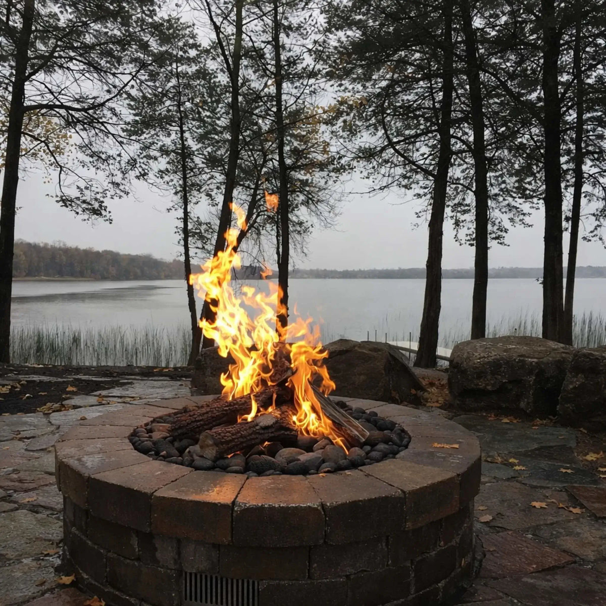 Custom circular fire pit with a sculptural metal globe centerpiece, surrounded by a ring of fire over black lava rock. The installation is set within a stone patio seating area with chairs and a scenic lake view in the background.