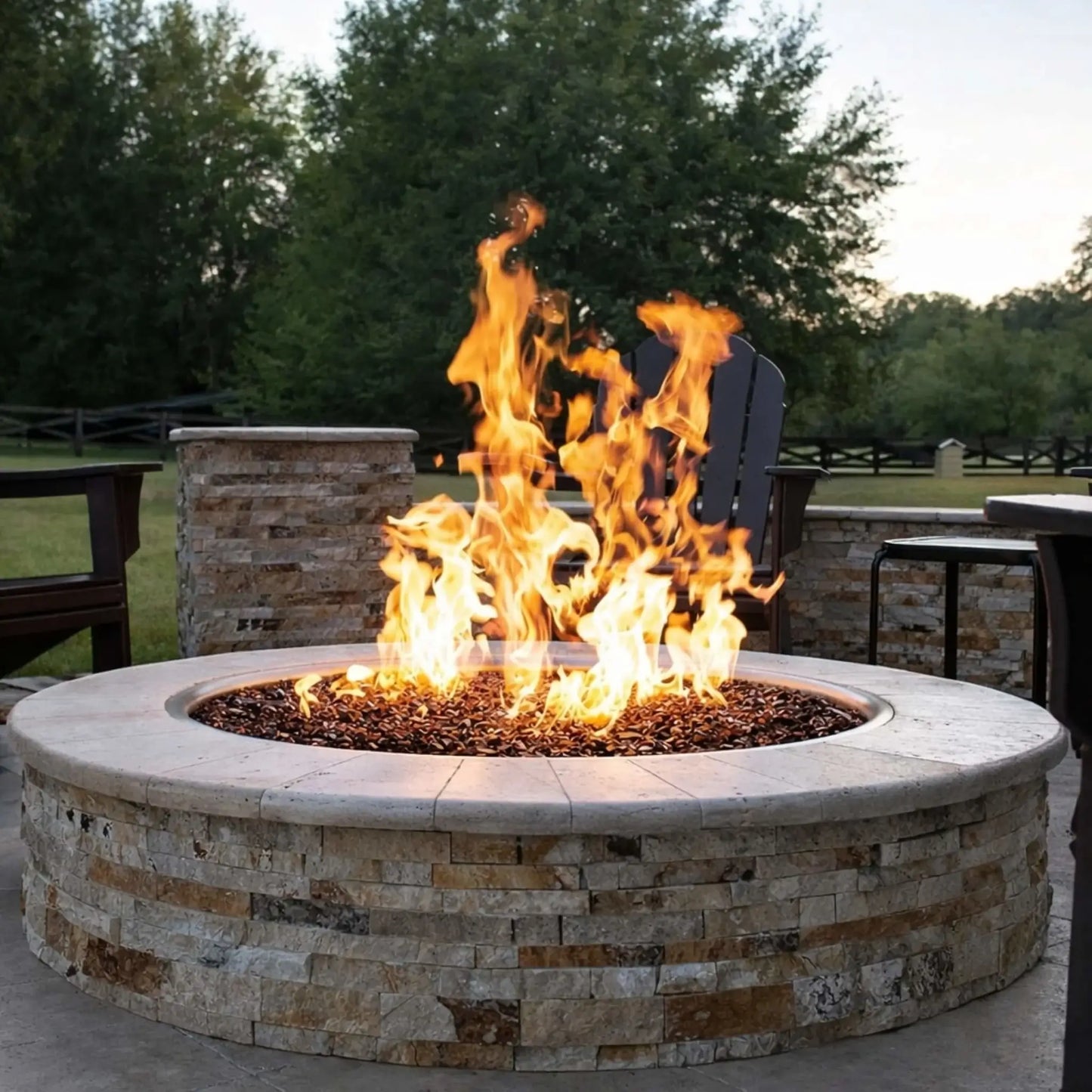 Stone-built round fire pit with a wide ledge and vibrant flames rising over decorative blue fire glass. Positioned on a concrete patio with outdoor seating, cushions, and backyard elements, this setup adds a colorful, contemporary focal point to the space.