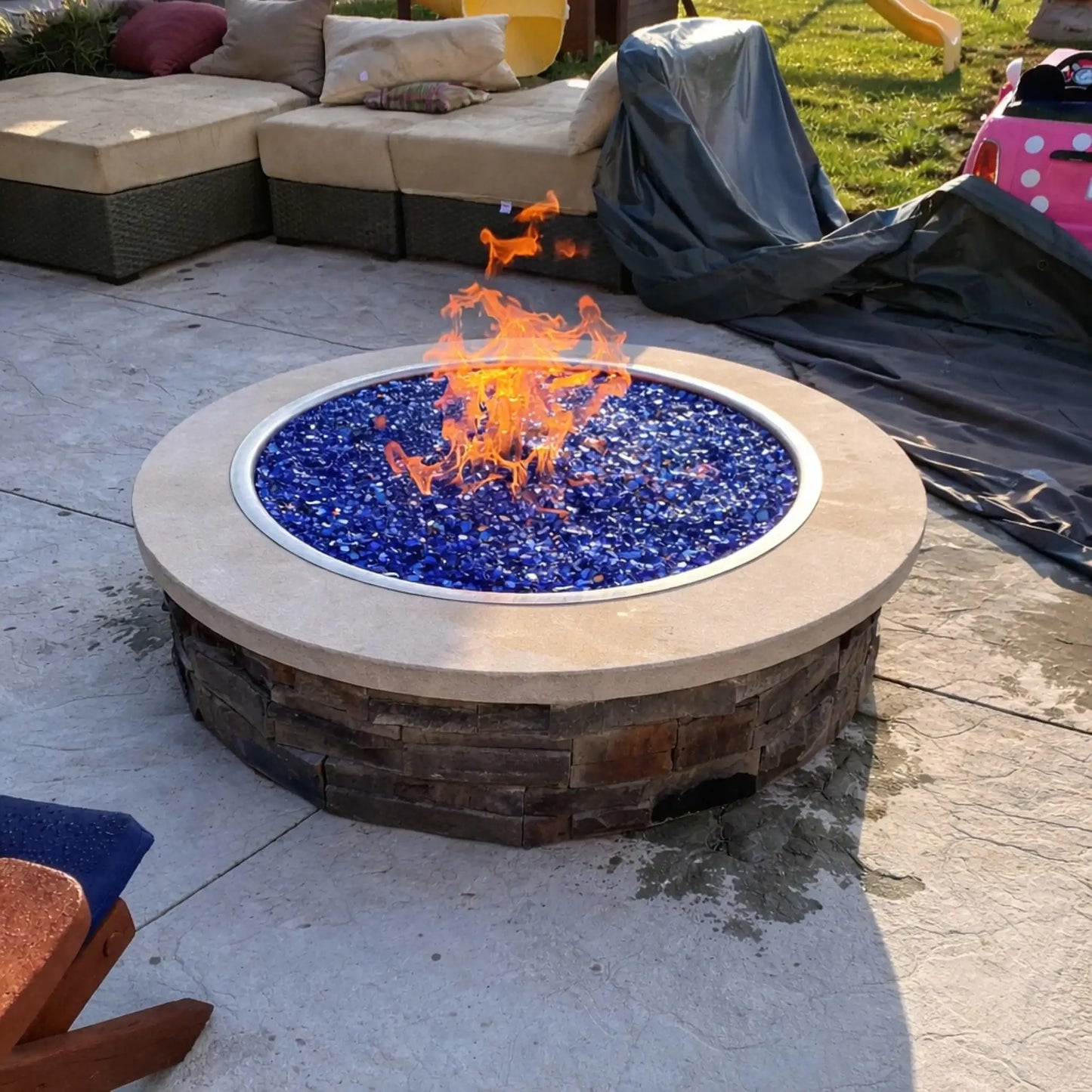 Large circular public fire pit surrounded by a metal safety railing, filled with glowing fire media and a central flame. Set under a covered pavilion with festive string lights and people gathered around, this scene captures a lively nighttime social atmosphere.