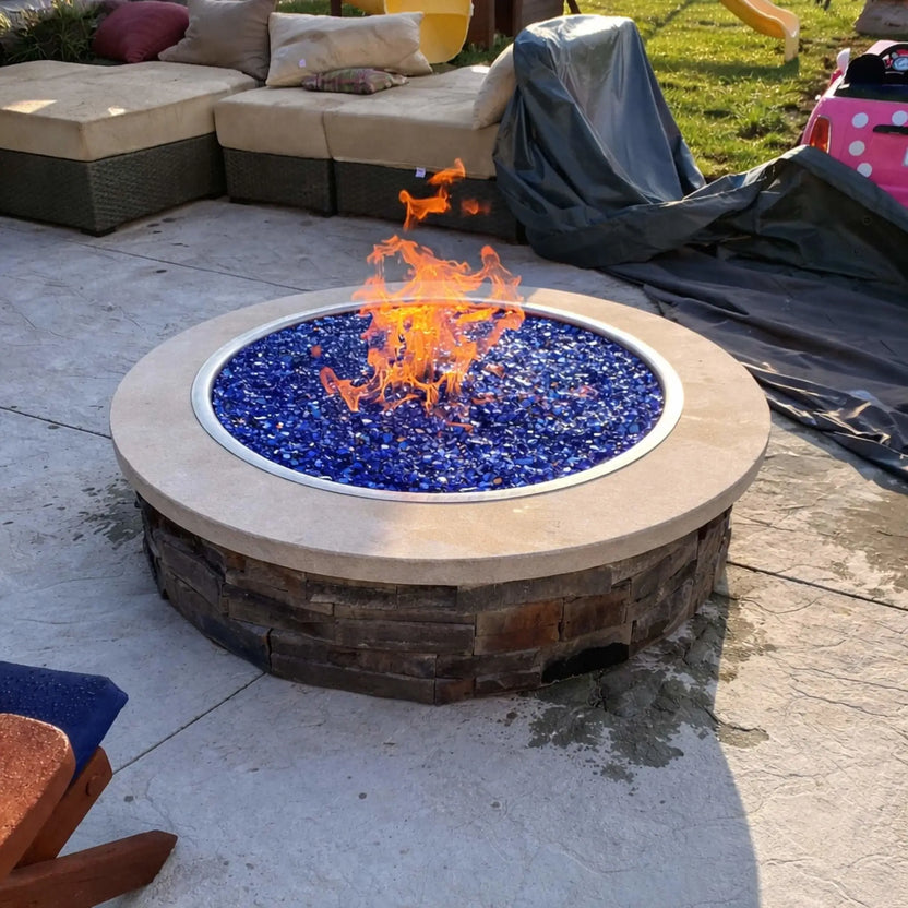 Large circular public fire pit surrounded by a metal safety railing, filled with glowing fire media and a central flame. Set under a covered pavilion with festive string lights and people gathered around, this scene captures a lively nighttime social atmosphere.
