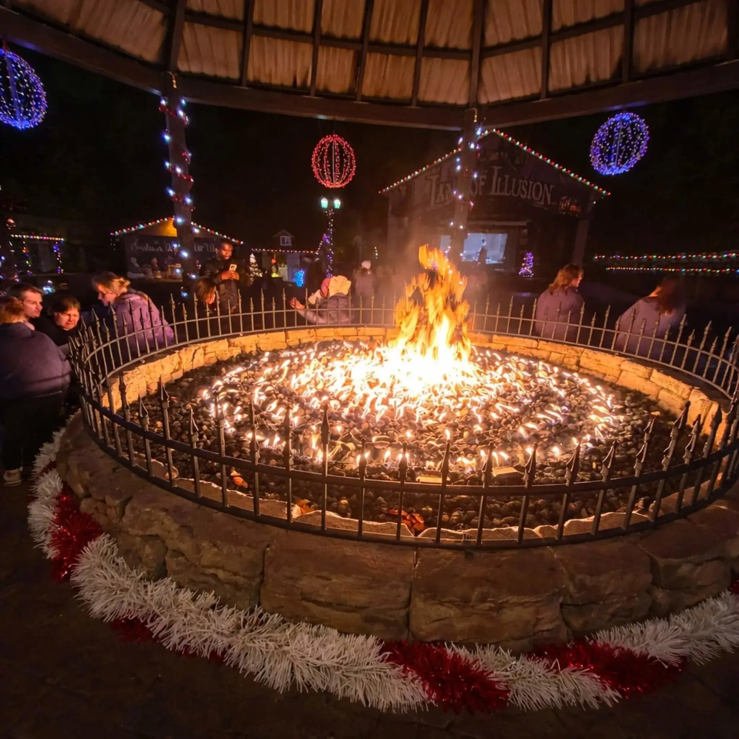 Traditional round stone fire pit with stacked logs burning over black lava rock, set within a paved patio area. Surrounded by a curved retaining wall, the design offers a cozy and timeless outdoor seating space for relaxing evenings.