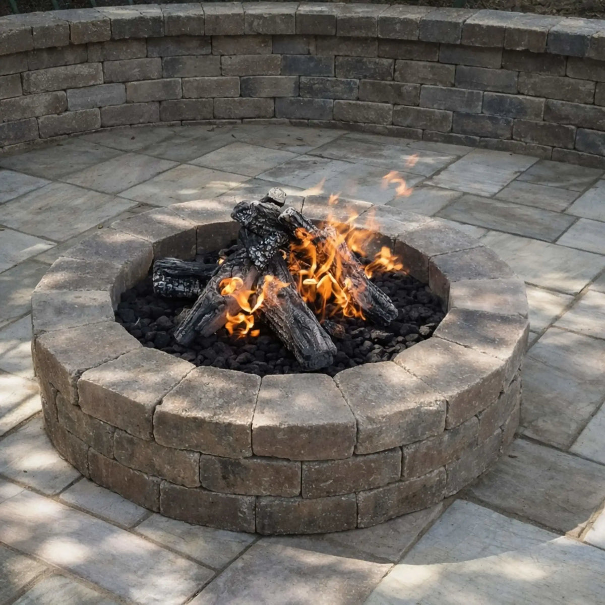 Traditional round stone fire pit with stacked logs burning over black lava rock, set within a paved patio area. Surrounded by a curved retaining wall, the design offers a cozy and timeless outdoor seating space for relaxing evenings.