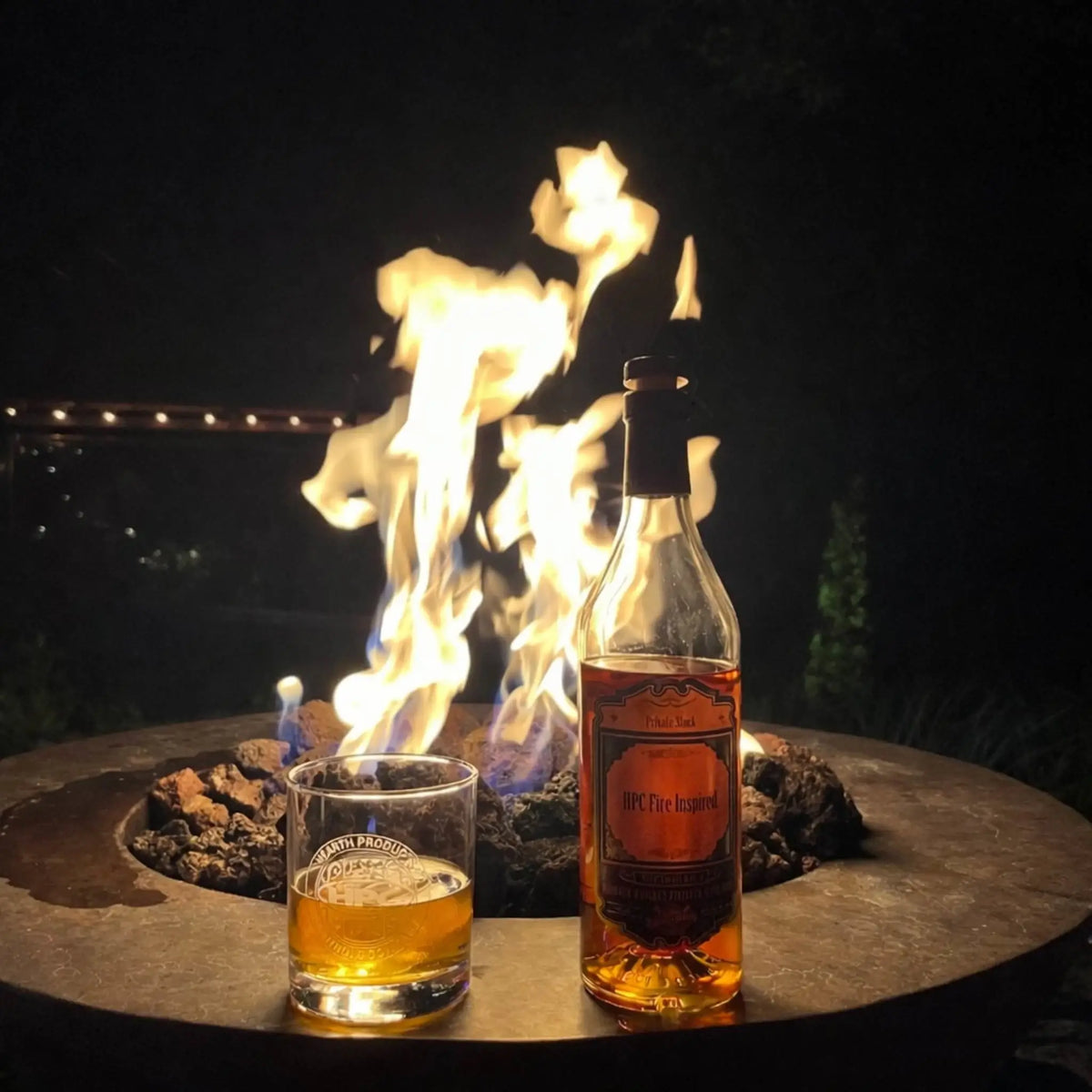 Nighttime patio fire pit scene featuring a metal fire table with lava rock and bright flames, accompanied by a glass of whiskey and a bottle labeled “HPC Fire Inspired.” The warm glow creates an intimate, relaxed setting ideal for evening enjoyment.