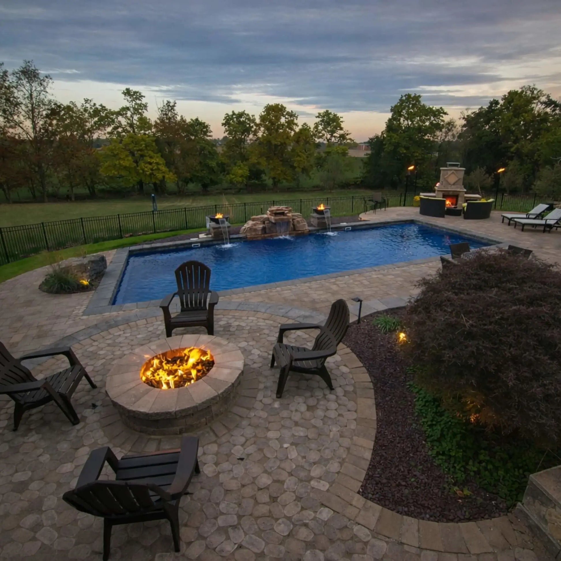 Luxury backyard with a circular gas fire pit surrounded by Adirondack chairs, overlooking a swimming pool and landscaped yard. The fire feature emits a steady flame, enhancing the ambiance of the outdoor living space during early evening hours.