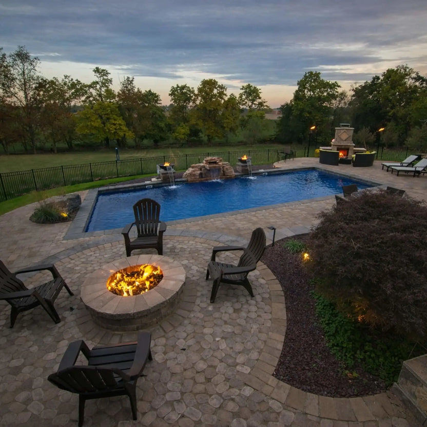 Luxury backyard with a circular gas fire pit surrounded by Adirondack chairs, overlooking a swimming pool and landscaped yard. The fire feature emits a steady flame, enhancing the ambiance of the outdoor living space during early evening hours.