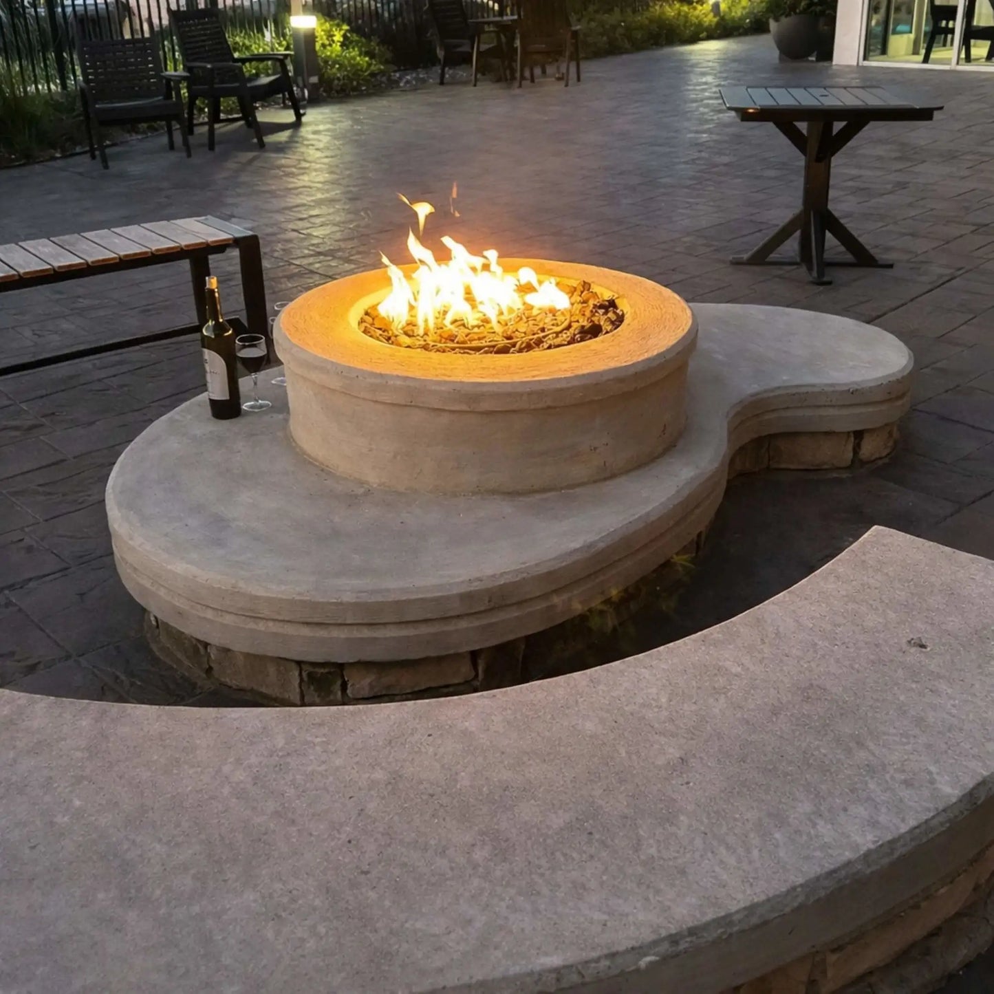 Curved concrete seating area integrated with a round gas fire pit, glowing with warm flames over decorative fire media. The surrounding patio includes outdoor tables, chairs, and ambient lighting, creating a comfortable social space for evening gatherings.