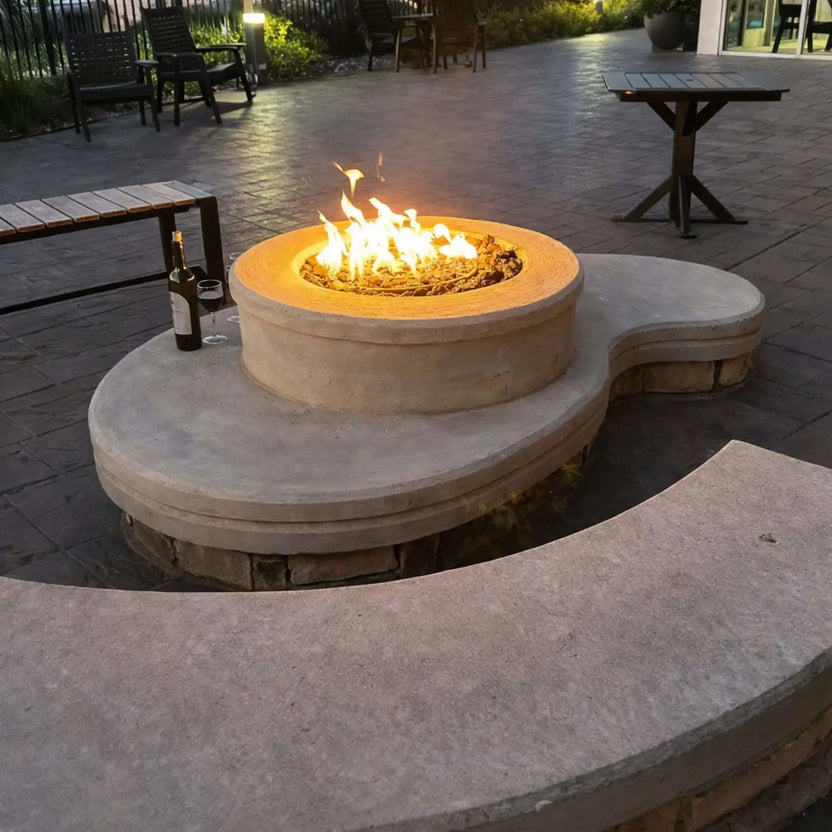 Curved concrete seating area integrated with a round gas fire pit, glowing with warm flames over decorative fire media. The surrounding patio includes outdoor tables, chairs, and ambient lighting, creating a comfortable social space for evening gatherings.