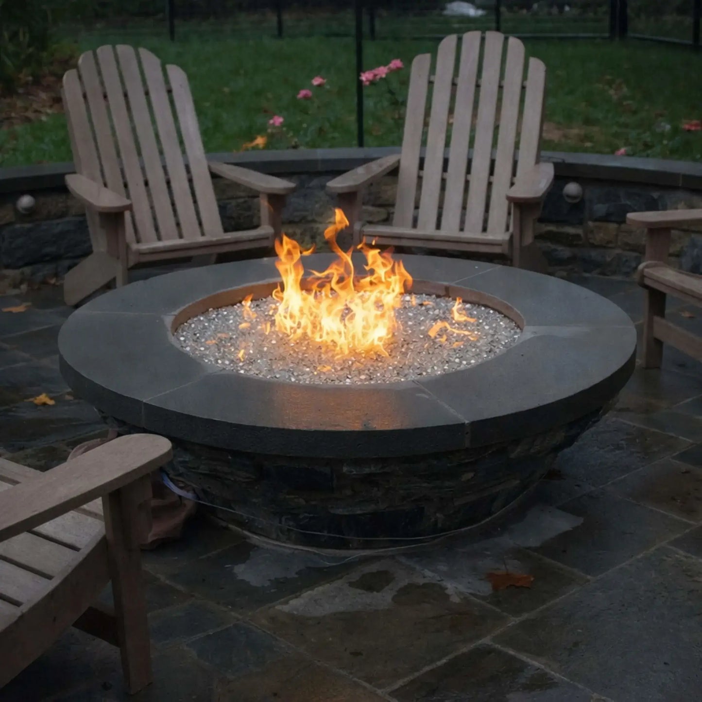 Modern circular fire pit with a smooth dark stone top and clear fire glass, surrounded by wooden Adirondack chairs on a wet patio surface. The flames rise steadily in the center, offering a cozy gathering space in a landscaped backyard with greenery and soft lighting.